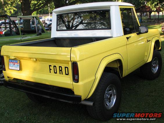 Yellow Ford Bronco with white top, rear view. Another green vehicle is parked nearby