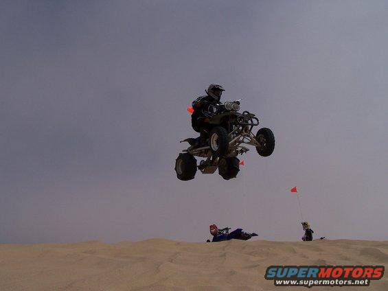 Quad bike rider airborne over sand dunes with two people on the ground