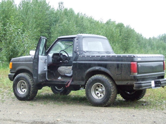 Black 1990 Ford Bronco with driver's door open and soft top installed, parked on a dirt road