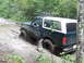 1990 Ford Bronco - An off-road vehicle navigates a challenging muddy path surrounded by dense foliage.