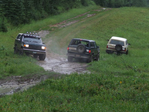 Three SUVs driving on a muddy, grassy hill; the lead vehicle is a dark green Ford Bronco from the late 1980s