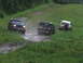 1990 Ford Bronco - Three SUVs navigate a muddy track on a grassy incline, with the lead vehicle being a dark green Ford Bronco.