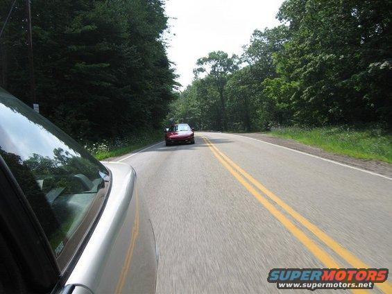 Silver car's front fender and windshield in foreground, red 1995 Ford Probe driving ahead on a two-lane road