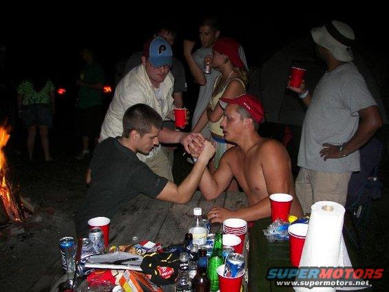 Group of people at a campfire, two men arm wrestling at a table with red cups and drinks
