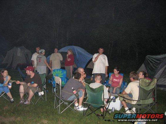 People gathered around a campfire at a campsite at night, with tents set up in the background