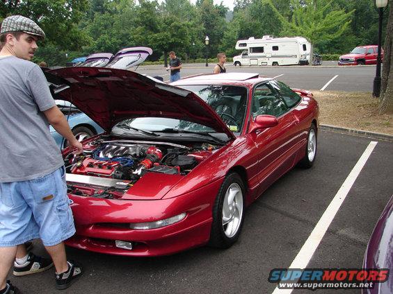 Red coupe with hood open, showing engine. A man in a cap and shorts stands nearby. Other vehicles and an RV are in the background