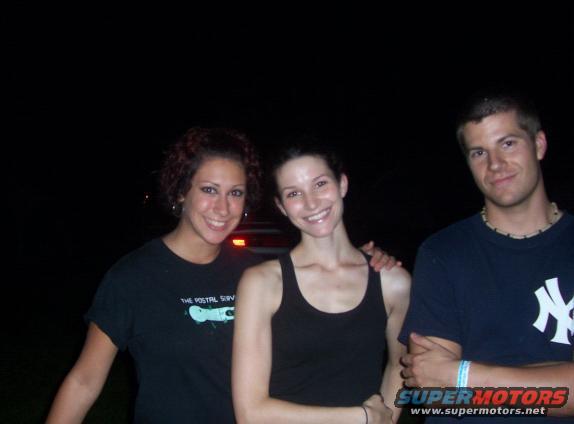 Three people smiling for a photo at night. The woman on the left wears a black t-shirt with text and a graphic