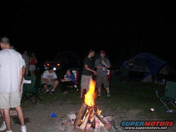 People gathered around a campfire at night with tents set up in the background
