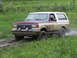 1990 Ford Bronco - A maroon and white Ford Bronco navigates a muddy, grassy terrain.