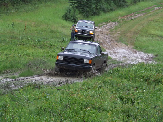 Black Ford F-150 pickup truck splashing through mud, followed by a dark Ford Bronco SUV on a dirt road