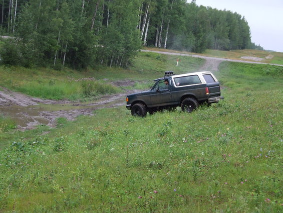 Dark green 1990 Ford Bronco with a white top drives on a grassy hill near a muddy track