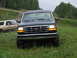 1990 Ford Bronco - Two Ford Broncos on a grassy, off-road path with trees in the background.