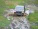 1990 Ford Bronco - A dark 1990 Ford Bronco navigates a muddy trail, kicking up water as it drives through a deep puddle.