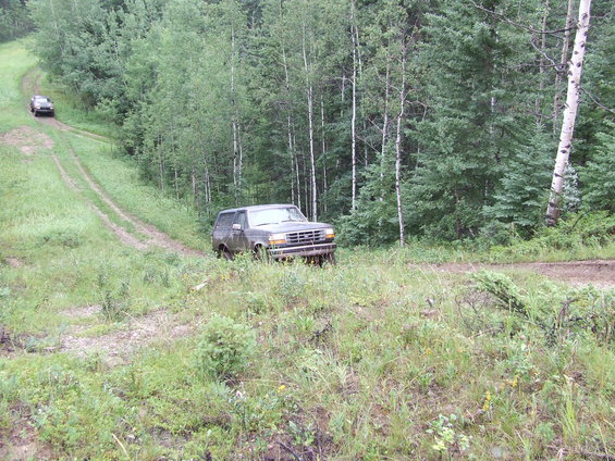 1990 Ford Bronco driving on a dirt road in a wooded area, another vehicle is visible further up the road