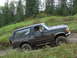 1990 Ford Bronco - A dark green 1990 Ford Bronco navigates a muddy off-road trail surrounded by trees.