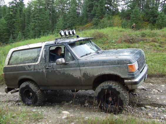 Muddy 1990s Ford Bronco with roof lights driving on a dirt trail