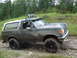1990 Ford Bronco - A woman drives a dirty Ford Bronco with off-road tires and roof lights through a wooded area.