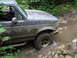1990 Ford Bronco - A mud-splattered 1990 Ford Bronco navigates a rocky stream in a wooded area.