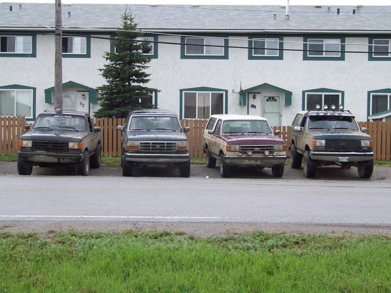 Four SUVs parked in a row in front of townhouses. The leftmost is black, followed by a dark blue, a maroon and white, and a green and tan one