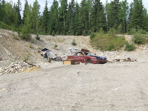 Two wrecked cars, one red and one black, in a gravelly junkyard with trees in the background
