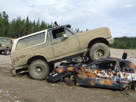 Muddy Ford Bronco on top of a rusted, wrecked car with graffiti. Another SUV is in the background