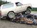 1990 Ford Bronco - A 1990 Ford Bronco rests atop a heavily damaged car in a junkyard setting.