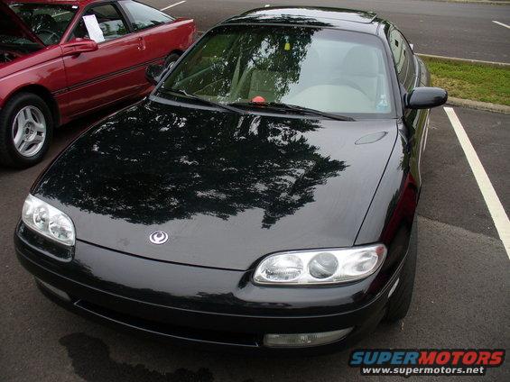 Black coupe front view with circular emblem. Red car in background