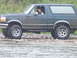 1990 Ford Bronco - A dark green 1990 Ford Bronco navigates a rocky stream bed, with a driver visible inside.