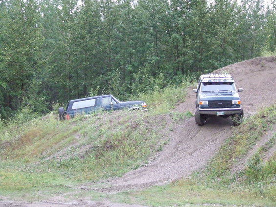 1990 Ford Bronco and another SUV on a dirt hill with trees in background