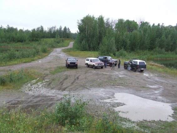 Four SUVs and trucks parked on a muddy dirt road with trees and a pond in the background