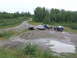 1990 Ford Bronco - A group of off-road vehicles are gathered on a dirt path near a body of water, surrounded by dense forest.