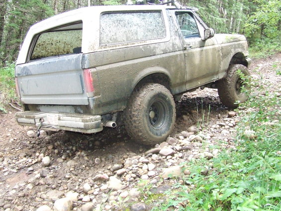 Mud-covered 1990 Ford Bronco driving on a rocky, wooded trail