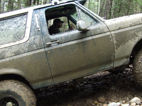 Mud-covered blue Ford Bronco, side view, driver visible, off-road terrain