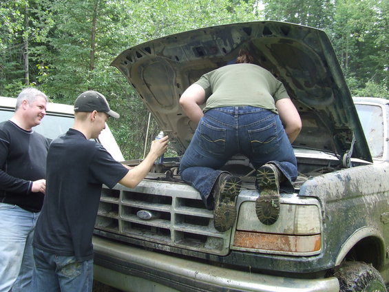 Grubby Ford Bronco with hood open, two men watching another work on engine