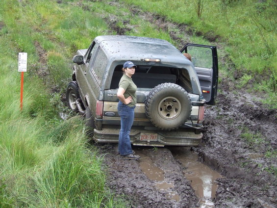 Woman standing next to a muddy Ford Bronco with its rear stuck in a mud pit