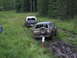 1990 Ford Bronco - Two Ford Broncos navigate a muddy off-road trail surrounded by trees.