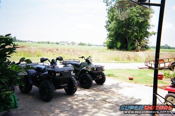 Two ATVs parked on a driveway, one black and one green. A field and trees are in the background