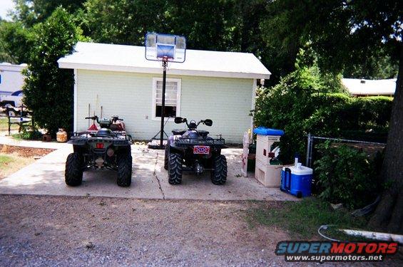 Two ATVs parked on a concrete driveway in front of a light green building with a basketball hoop