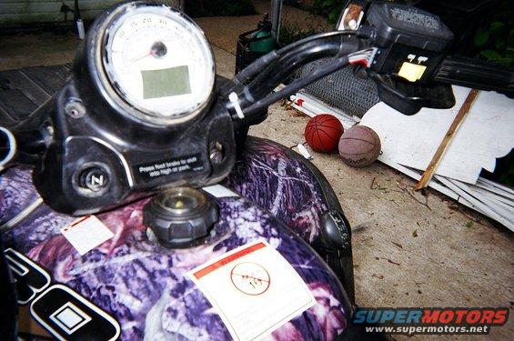 ATV dashboard with gauge, handlebars, and fuel cap; two basketballs are nearby