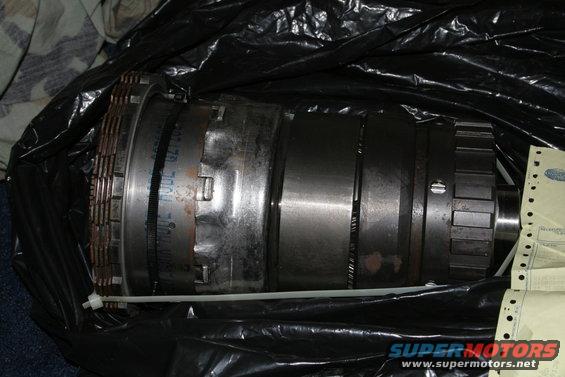 Close-up of a transmission part, possibly a torque converter, wrapped in black plastic. Some text is visible on the part