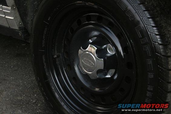 Black steel wheel with a chrome Ford hubcap and a black tire. The side panel of the vehicle is visible