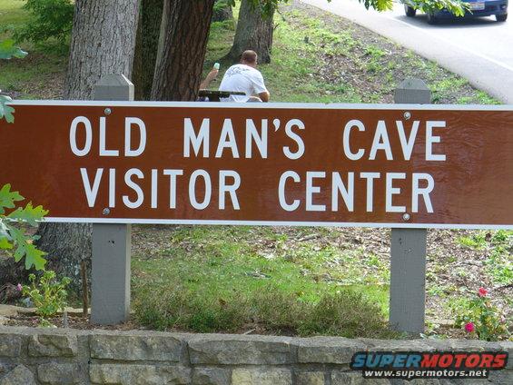 Brown sign reading "OLD MAN'S CAVE VISITOR CENTER" with a person sitting behind it