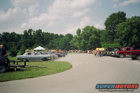 Outdoor car show with rows of cars and trucks parked, people milling around, and trees in the background