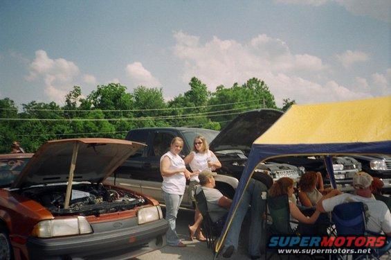 Orange car with hood open, front view. People stand nearby under a yellow tent