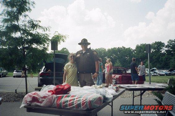 People gathered outdoors, some standing near a table with wrapped items. Several cars are parked in the background