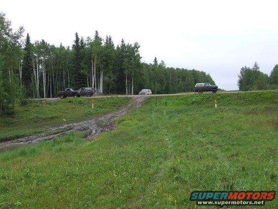 three SUVs driving on a dirt road next to a forest, one vehicle is on a muddy track