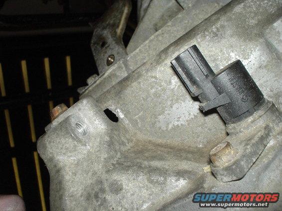Close-up of a gray metal automotive transmission housing with a black sensor attached. A small hole is visible on the housing