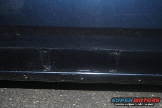 Close-up of a dark blue car's lower body panel showing some rivets and a slight dent