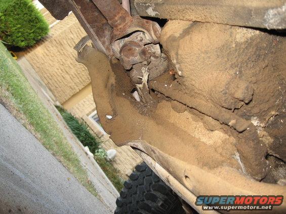 Underside of a dirty vehicle frame with visible drivetrain components and a tire tread