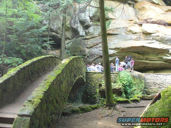 stone bridge in a wooded area with a rock cliff and people gathered